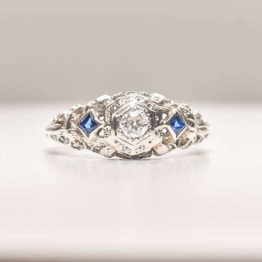 Art Deco 18K white gold engagement ring with diamond center and synthetic sapphire accents, size 6.25, with intricate filigree design.