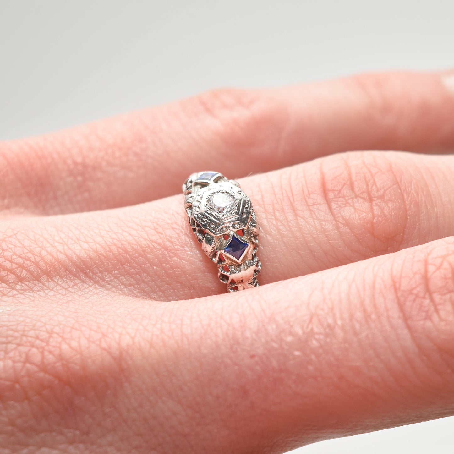 Art Deco 18K white gold diamond and sapphire filigree engagement ring with natural diamonds and synthetic sapphires, size 6.25 US, on a finger against a white background.