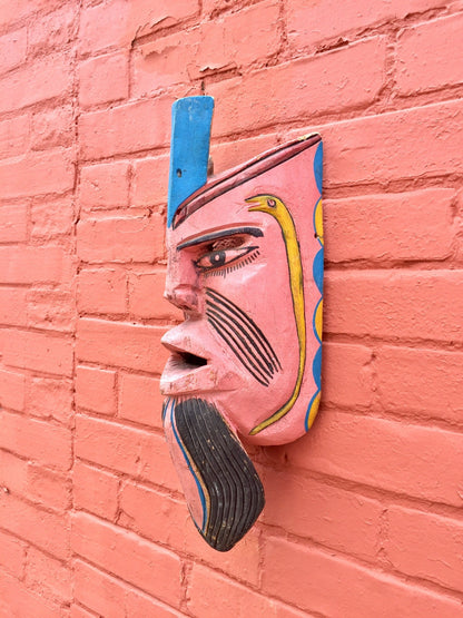 Colorful wooden mask hanging on pink brick wall featuring intricate carvings and designs in blue, pink, and yellow paint, creating an eye-catching vintage home decor or wall hanging piece.