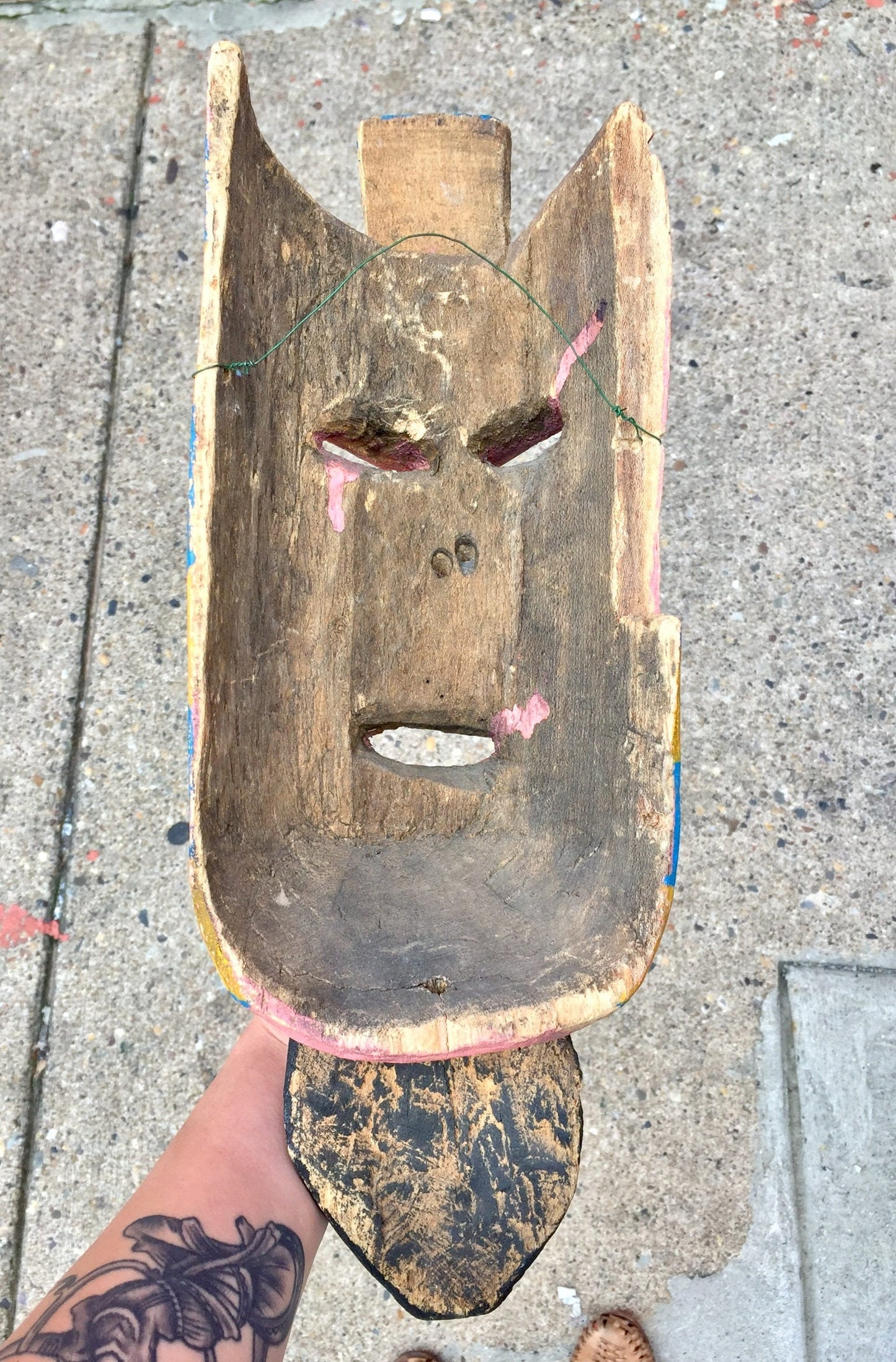 Vintage hand-carved wooden mask with painted details, mounted on concrete, alongside a person's tattooed arm and leg.