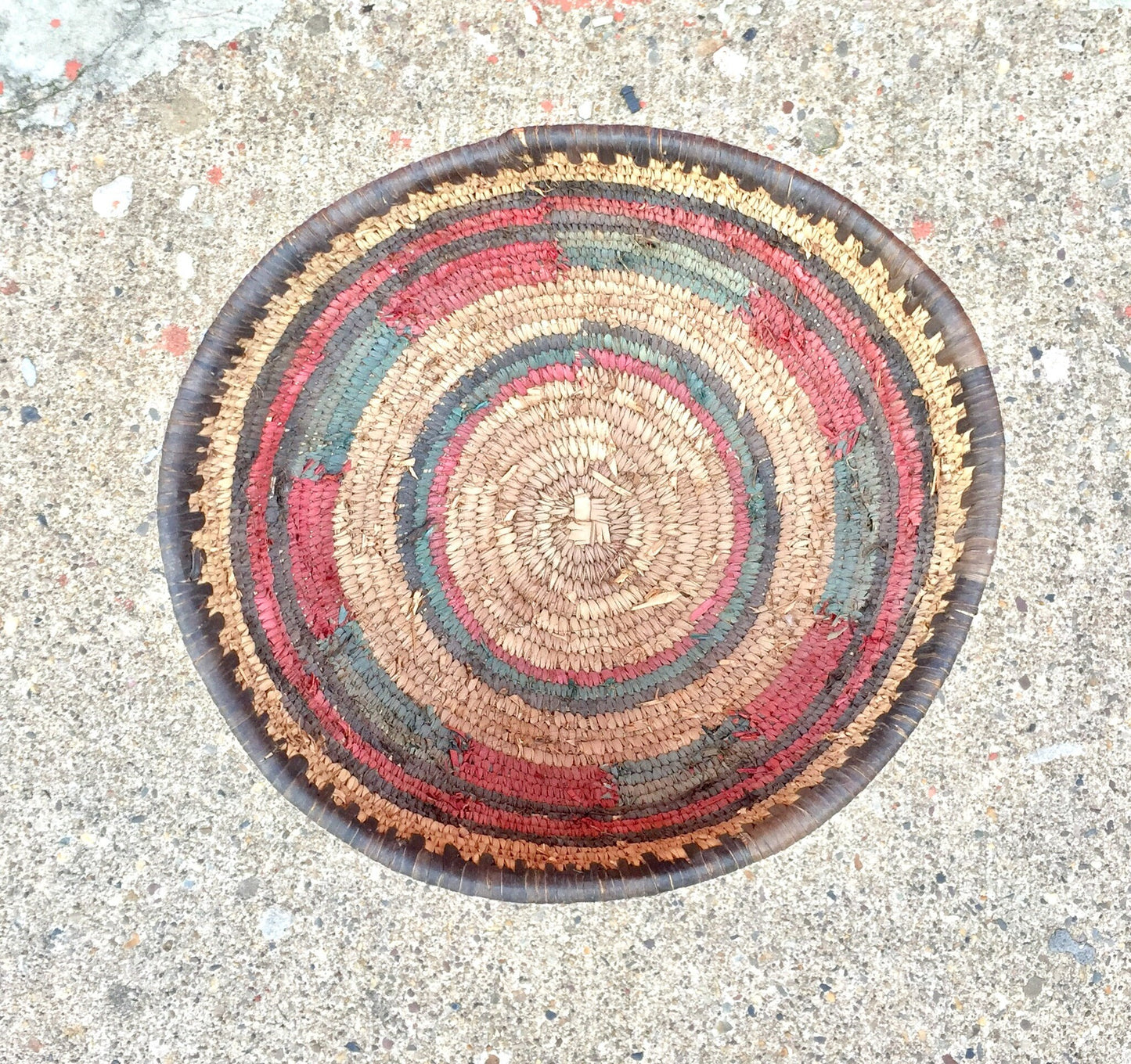 Round woven basket with colorful concentric rings in shades of red, orange, yellow, green and blue on a concrete surface