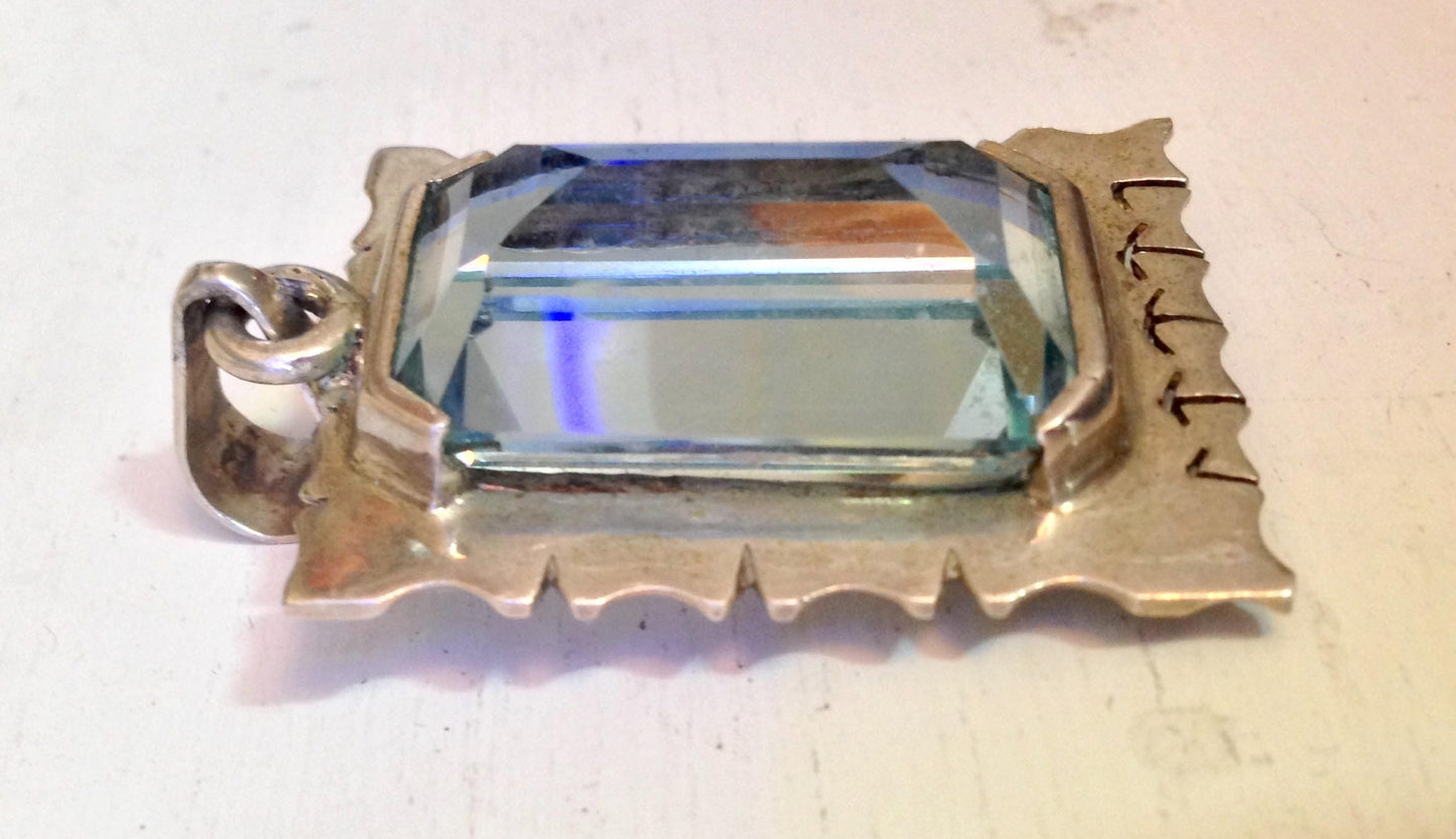 Vintage sterling silver pendant featuring a rectangular faceted aquamarine gemstone, with ornate silver detailing resembling a scroll or ribbon border.