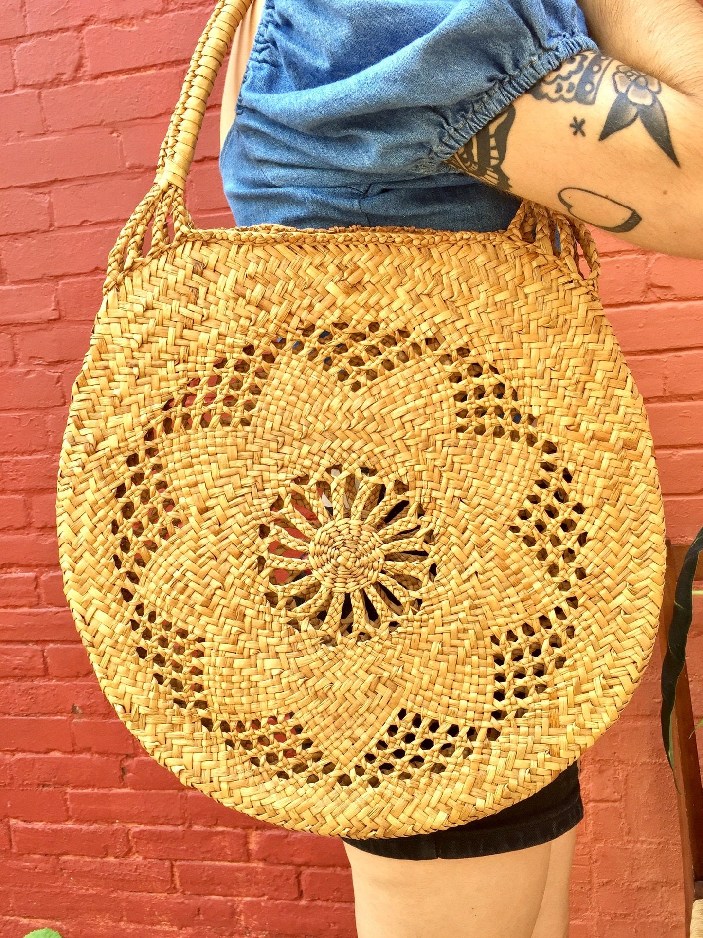 Round woven straw bag with intricate floral pattern design hanging on a person's tattooed arm against a red brick wall background.