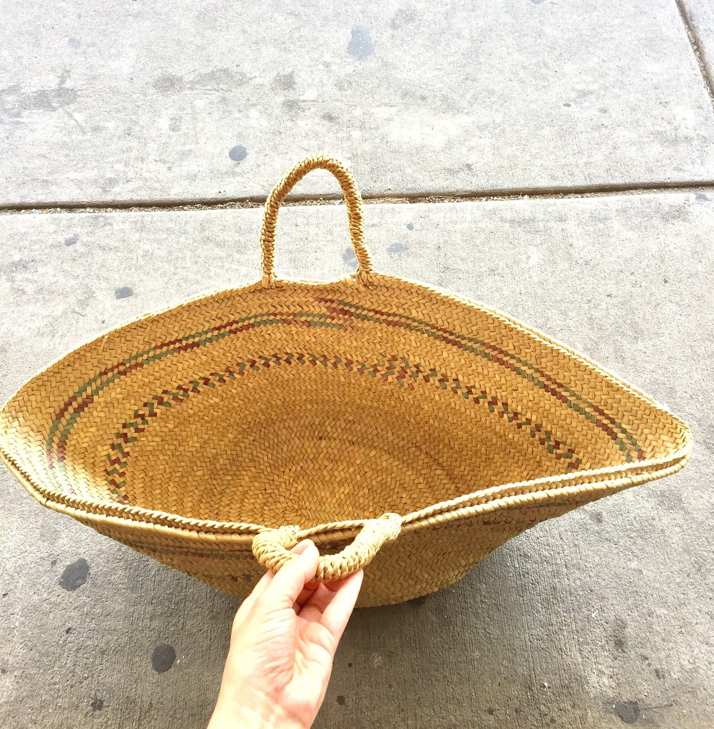 Hand holding a large handwoven straw tote bag with handles against a concrete background, boho style beach or shopping bag
