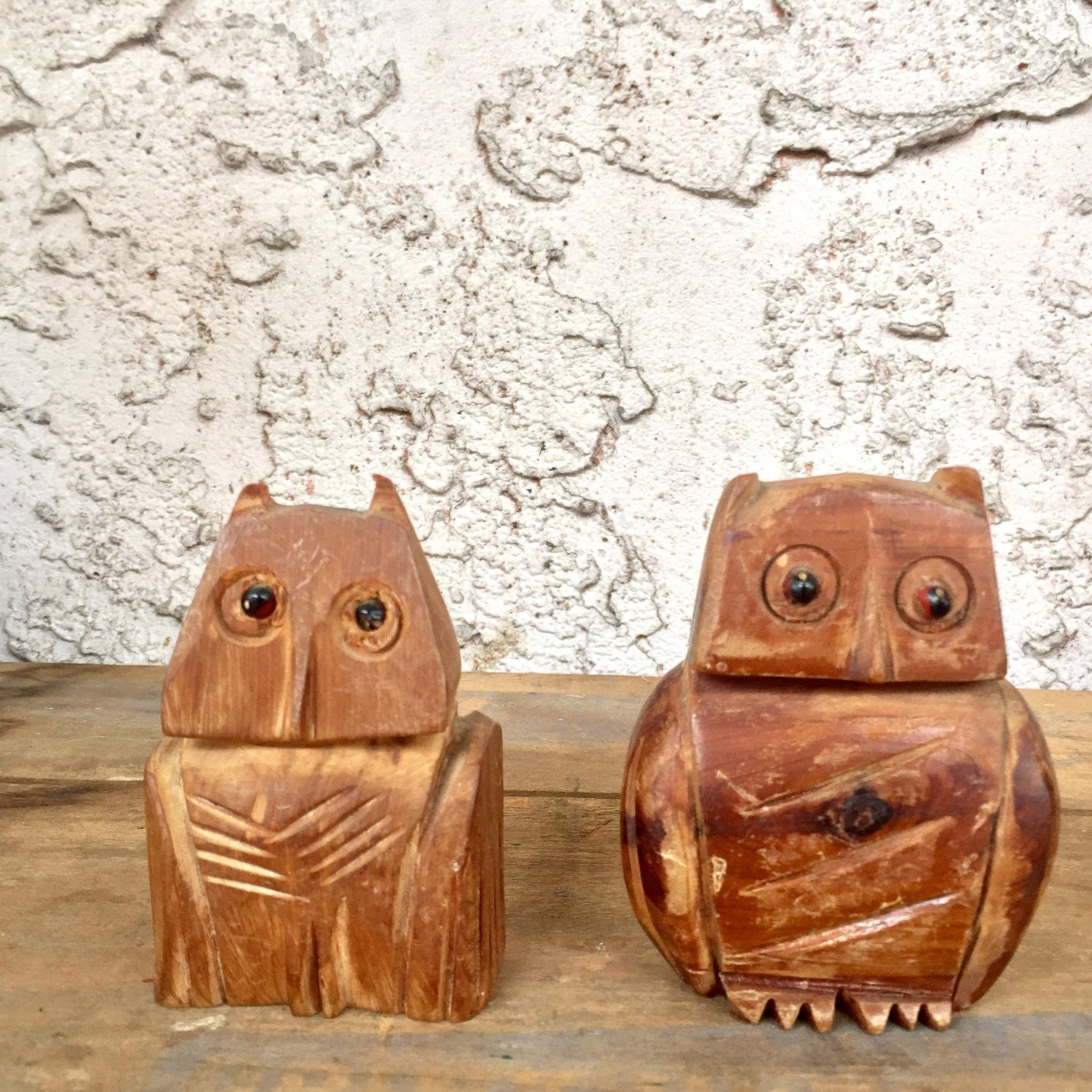 Pair of vintage hand-carved wooden owl figurines, one seated and one standing, with intricate textured details, against a textured white concrete wall background, suitable for use as unique pencil holders or desk organizers.