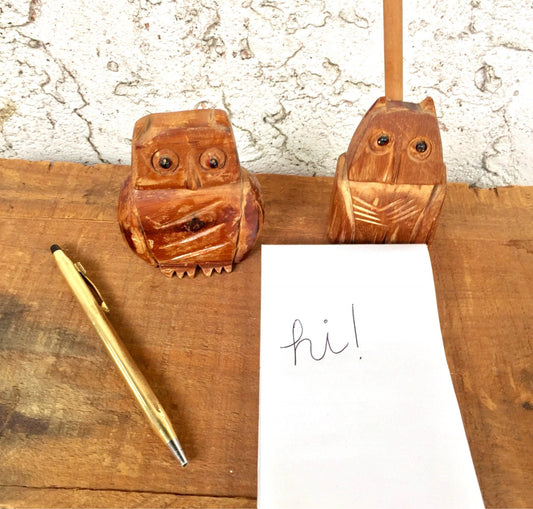 Two carved wooden owl figurines on a wooden surface, one with a note that says "hi!", next to a gold pen, suggesting a desk organizer or cute vintage folk art gift idea.