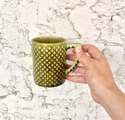 Vintage green ceramic mug with diamond pattern held in front of white textured wall