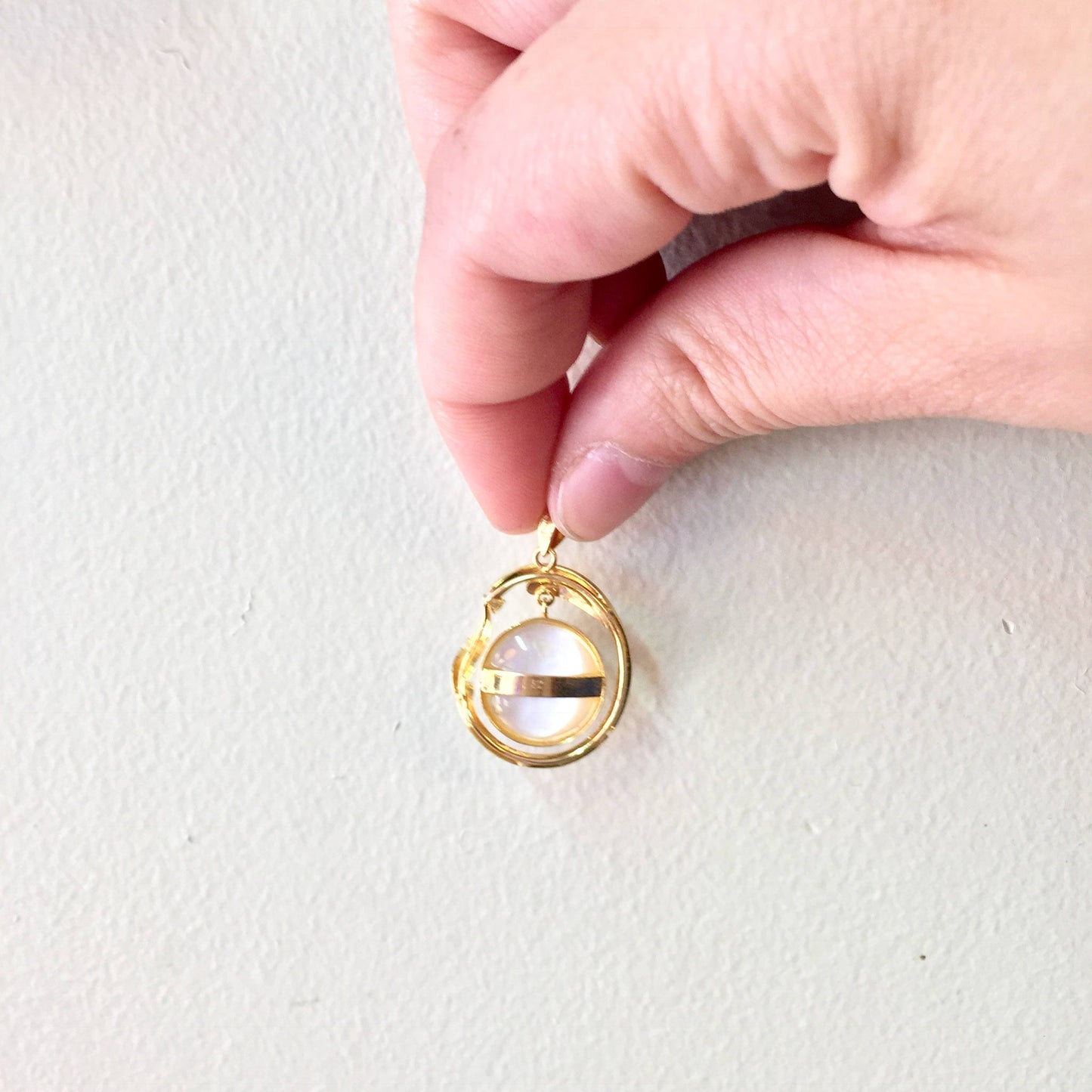 A hand holding a gold vintage-style pendant with a round pearl accent, against a white background.