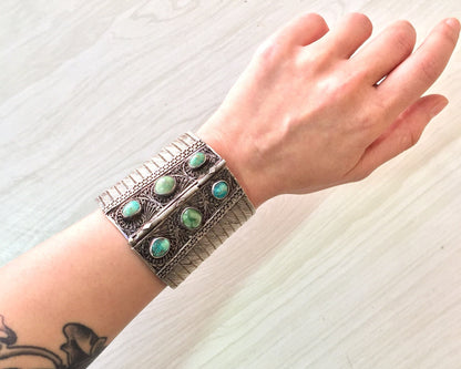 Vintage sterling silver cuff bracelet with intricate filigree design and inlaid turquoise cabochons, worn on a woman's wrist against a light textured background.