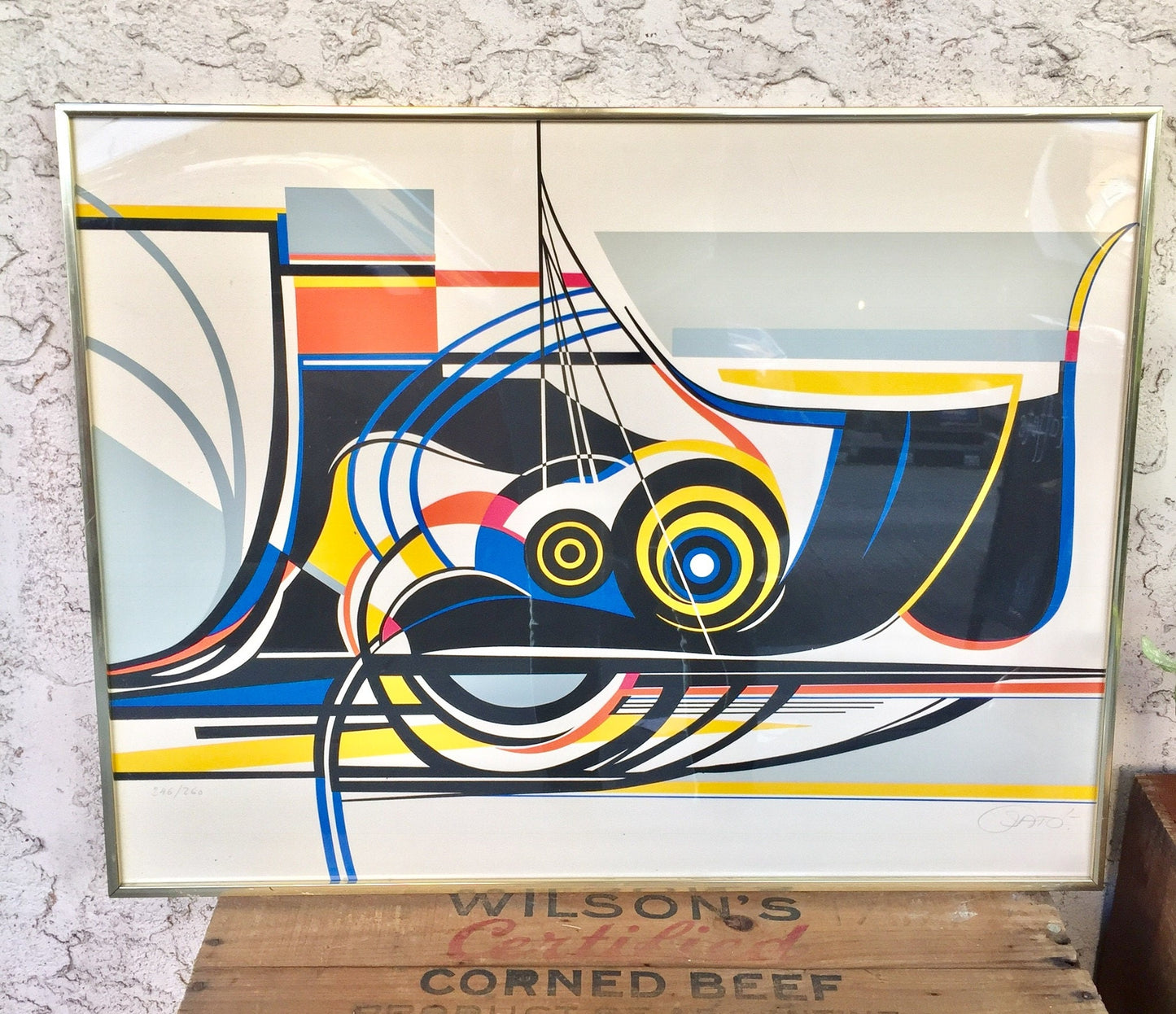 Abstract geometric artwork featuring overlapping circular and curved shapes in blue, red, yellow, black and white, resembling a stylized mechanical object or vehicle, signed by the artist Sato, displayed in a frame on a wooden surface against a textured white background.