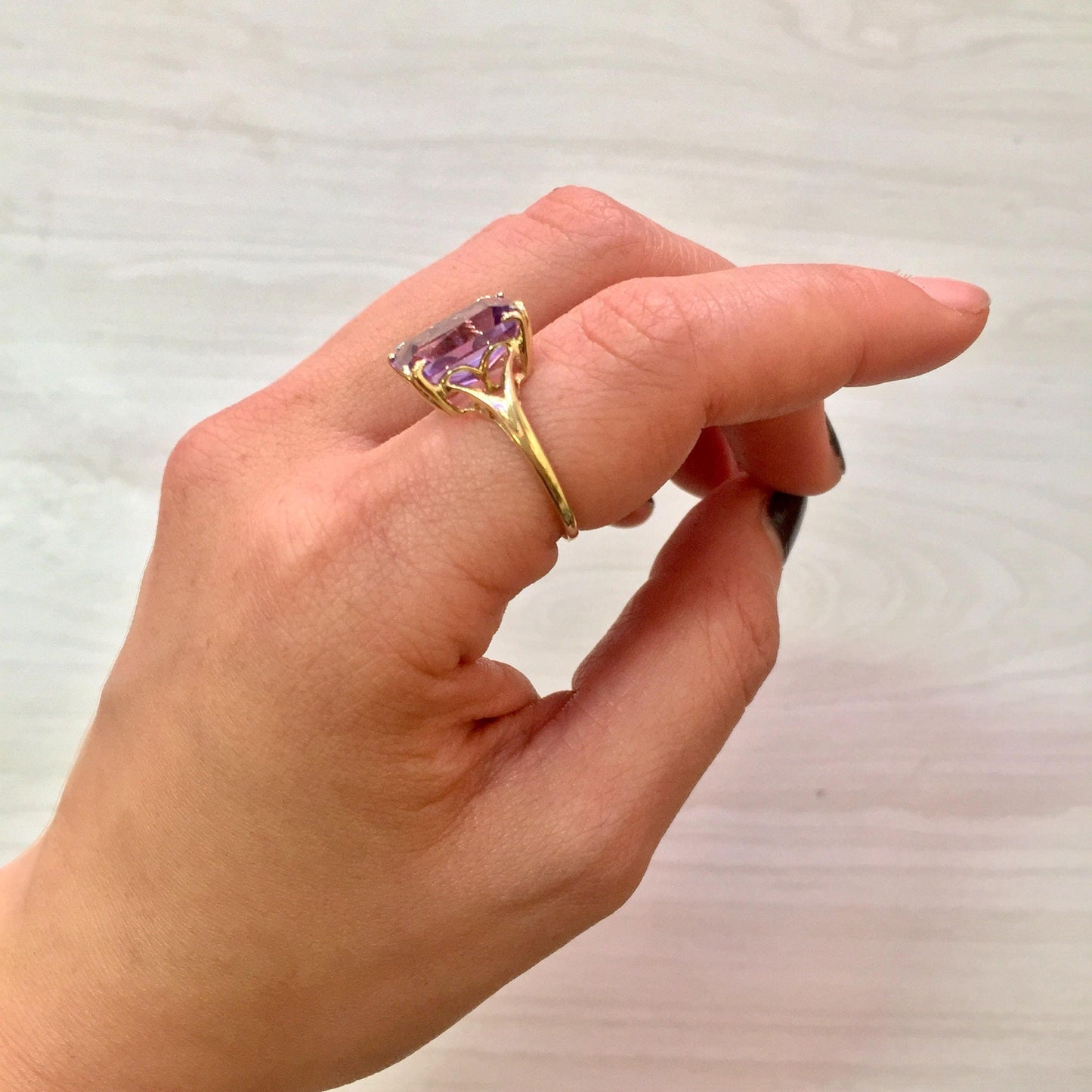 An emerald cut amethyst gemstone set in a 14 karat yellow gold ring, held between a person's fingers showcasing the ring's elegant design and vibrant purple color of the amethyst.