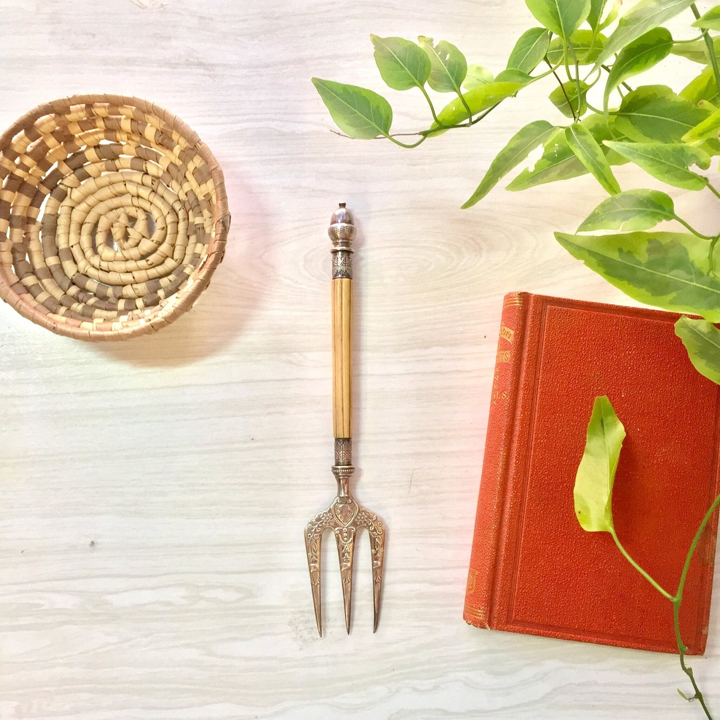 Antique Victorian bread fork with engraved bone handle, displayed next to a woven basket and book, surrounded by greenery on a textured background.