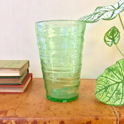 Vintage vaseline glass cup with rippled texture on a wooden table with books and plant illustrations in the background, hand-blown depression-era collectible glassware with pontil mark.