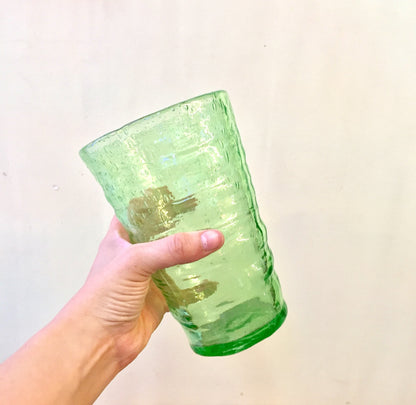 Hand holding a large, vintage green vaseline glass cup with a rippled texture, likely hand-blown during the Depression era. The glass has a pontil mark on the bottom, indicating it is a collectible piece.