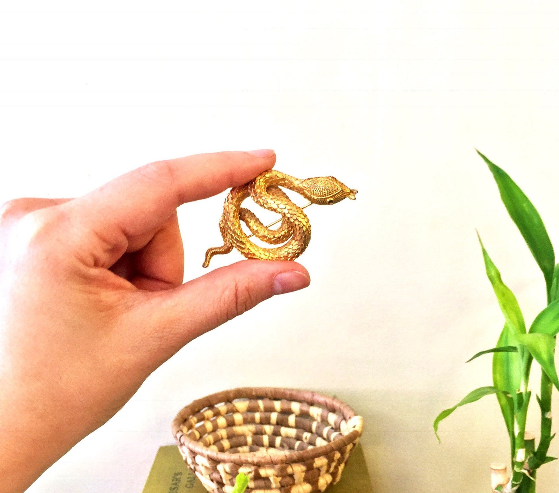 Gold tone coiled snake brooch pin held in hand above woven basket, next to green plant against white background, vintage costume jewelry gift idea