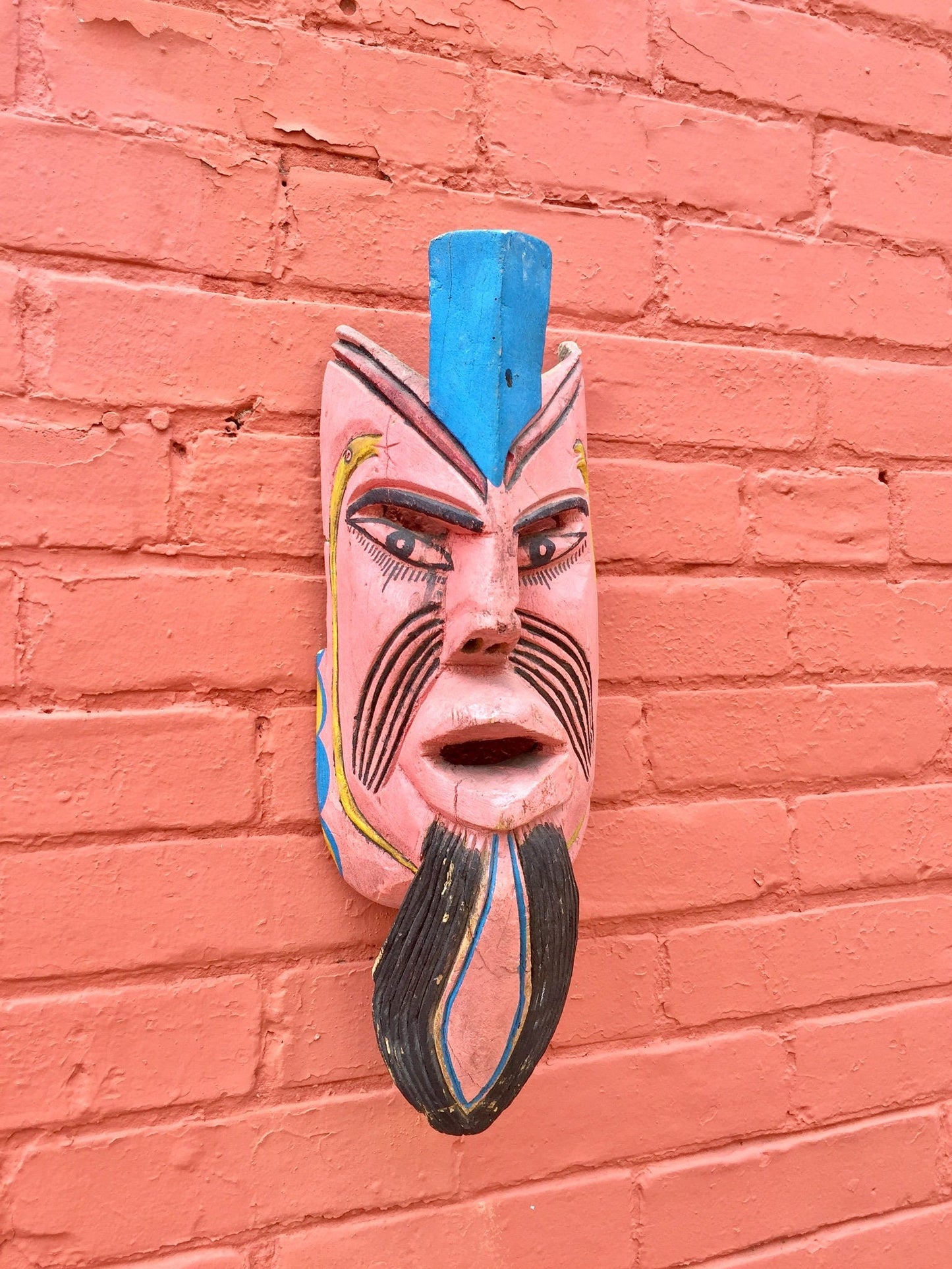 Colorful wooden mask with geometric designs hanging on a textured red brick wall.