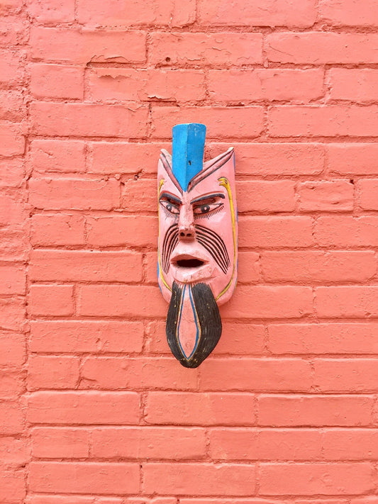 Vintage hand-carved wooden mask with blue, red and white colors mounted on a salmon-colored brick wall, adding an ethnic and decorative touch to the home decor.