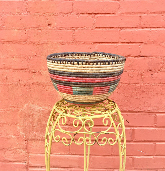 Colorful woven basket on yellow metal stand against pink brick wall, handmade bohemian vintage home decor