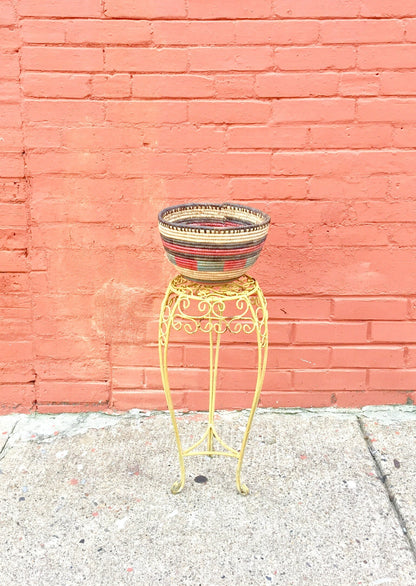 Colorful woven basket on ornate metal stand against pink brick wall, boho vintage home decor