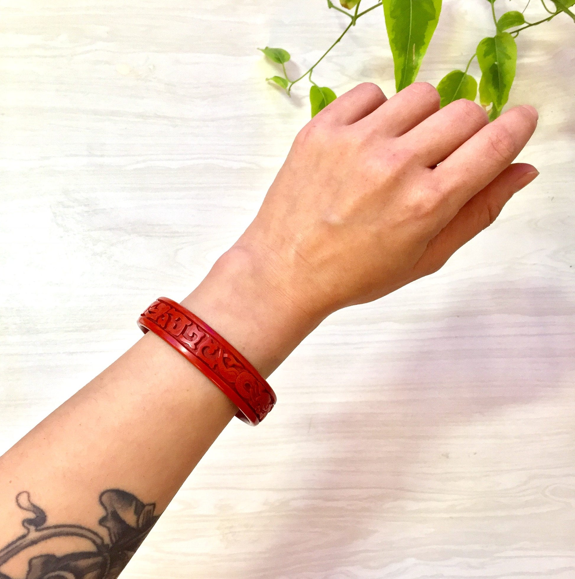 Red carved cinnabar bangle bracelet with dragon motif worn on tattooed wrist, held up against white textured background with green plant leaves at top of image.