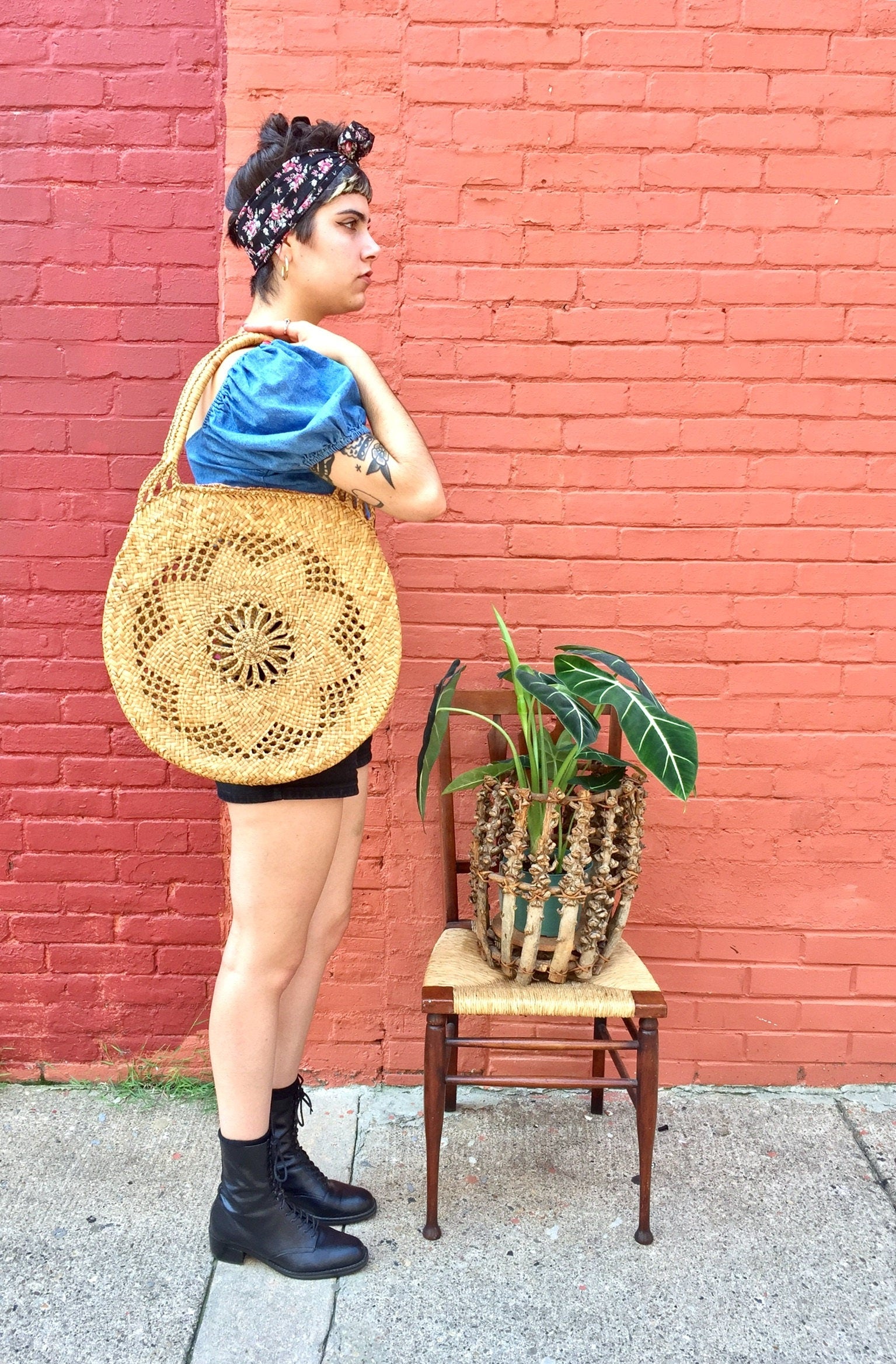Stylish woman wearing a blue top, patterned yellow tote bag, and black boots standing next to a potted plant in front of a brick wall.