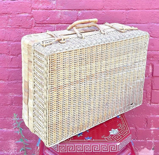 Vintage 1960's wicker briefcase with rattan construction, suitable for picnics, storage, crafts or rustic boho decor. The rectangular case has a hinged lid with two leather handle straps and sits atop a colorful patterned cloth against a bright pink brick wall background.
