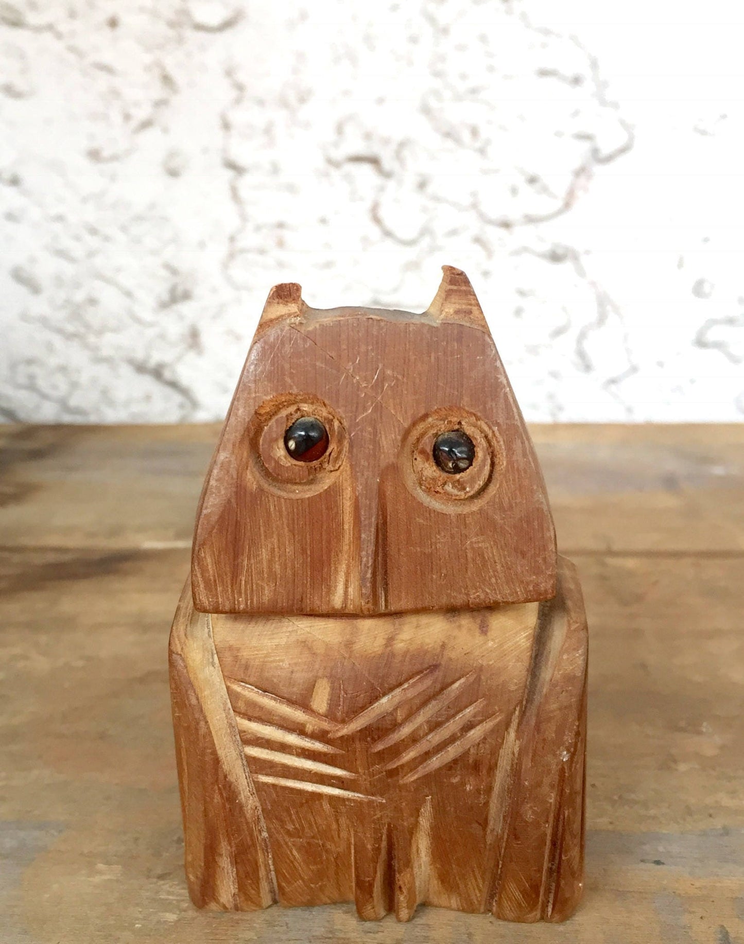 Wooden owl pencil holder carved in folk art style, with large eyes and textured feather details, sitting on a wooden surface against a white wall background.