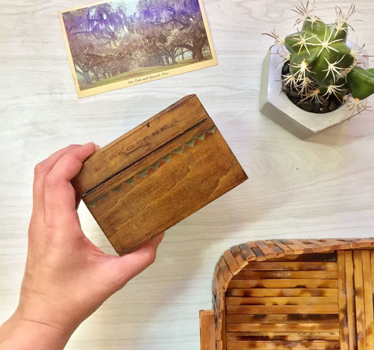 Vintage wooden box with intricate hand-carved details, held open to reveal natural wood grain interior. Shown alongside a small succulent plant and old sepia-toned photograph depicting a forest setting.