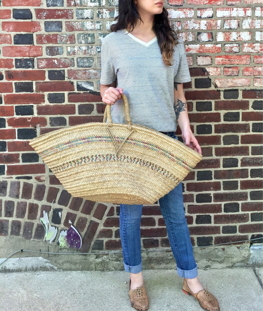 Natural woven straw tote bag held by a woman wearing a striped shirt and jeans, standing in front of a brick wall. The large, handmade boho-style beach bag features intricate woven patterns and handles.