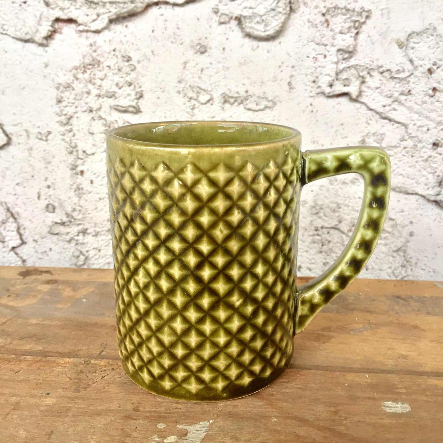 Vintage green ceramic mug with textured diamond pattern on a wooden surface against a weathered white brick wall background.