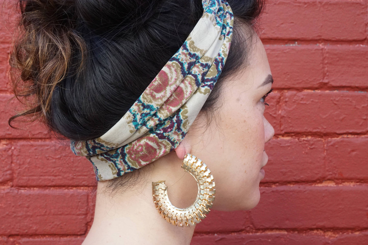 Closeup of a woman wearing a colorful floral headband and large gold hoop earrings, posing in front of a red brick wall