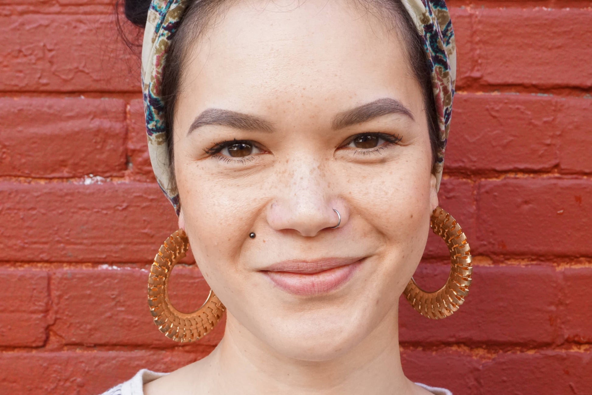 Close-up portrait of a smiling young woman with freckles wearing a floral head scarf and large gold hoop earrings against a red brick wall background.