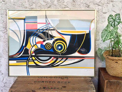 Modern abstract lithograph print featuring geometric shapes and lines in bright colors including yellow, blue, black and red, signed by the artist Sato. The framed artwork is displayed on a textured white wall next to a potted plant.