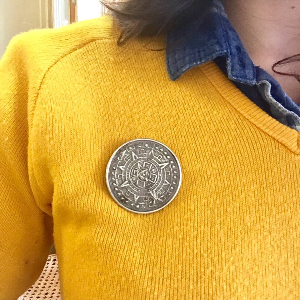 Vintage sterling silver Mayan Aztec sun calendar pendant brooch pinned on a yellow knit sweater. The circular pendant has an intricate carved pattern representing an ancient sundial calendar. The sweater collar is denim blue, adding a pop of color contrast to the bright yellow knit material.