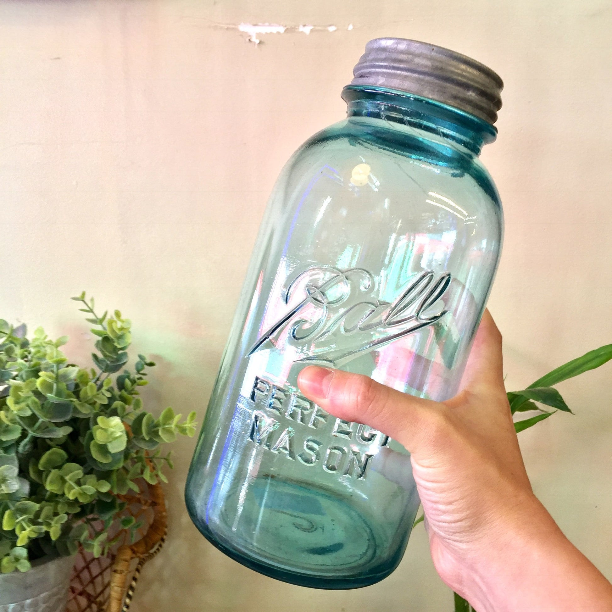 Antique blue hand-blown Ball Mason canning jar with zinc lid held in hand against neutral background with greenery.