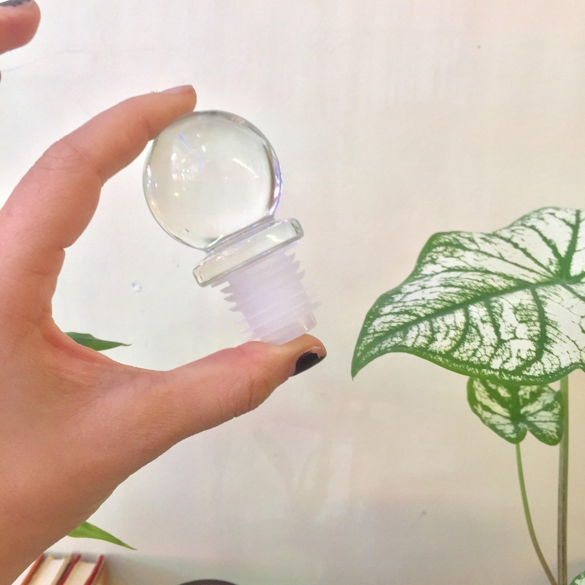 A hand holding a small clear glass bottle with a round stopper near a green leaf plant against a plain background.