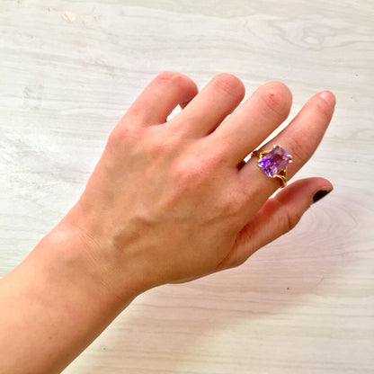 A hand wearing a 14 karat gold ring with a large emerald cut amethyst gemstone against a light wooden background.