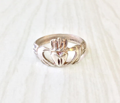 Sterling silver Claddagh ring with carved heart held by two hands, representing love, loyalty and friendship in Irish tradition, on white fabric background.