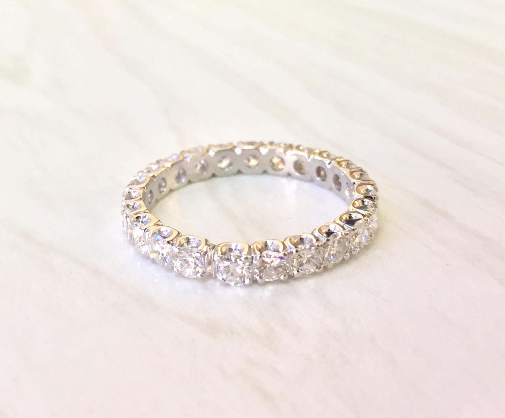 14 karat white gold eternity wedding band ring with round brilliant cut diamonds set all the way around the band. The diamonds are prong set with a scalloped edge design on the ring. The ring has a bright polish finish and sits on a light colored fabric background.