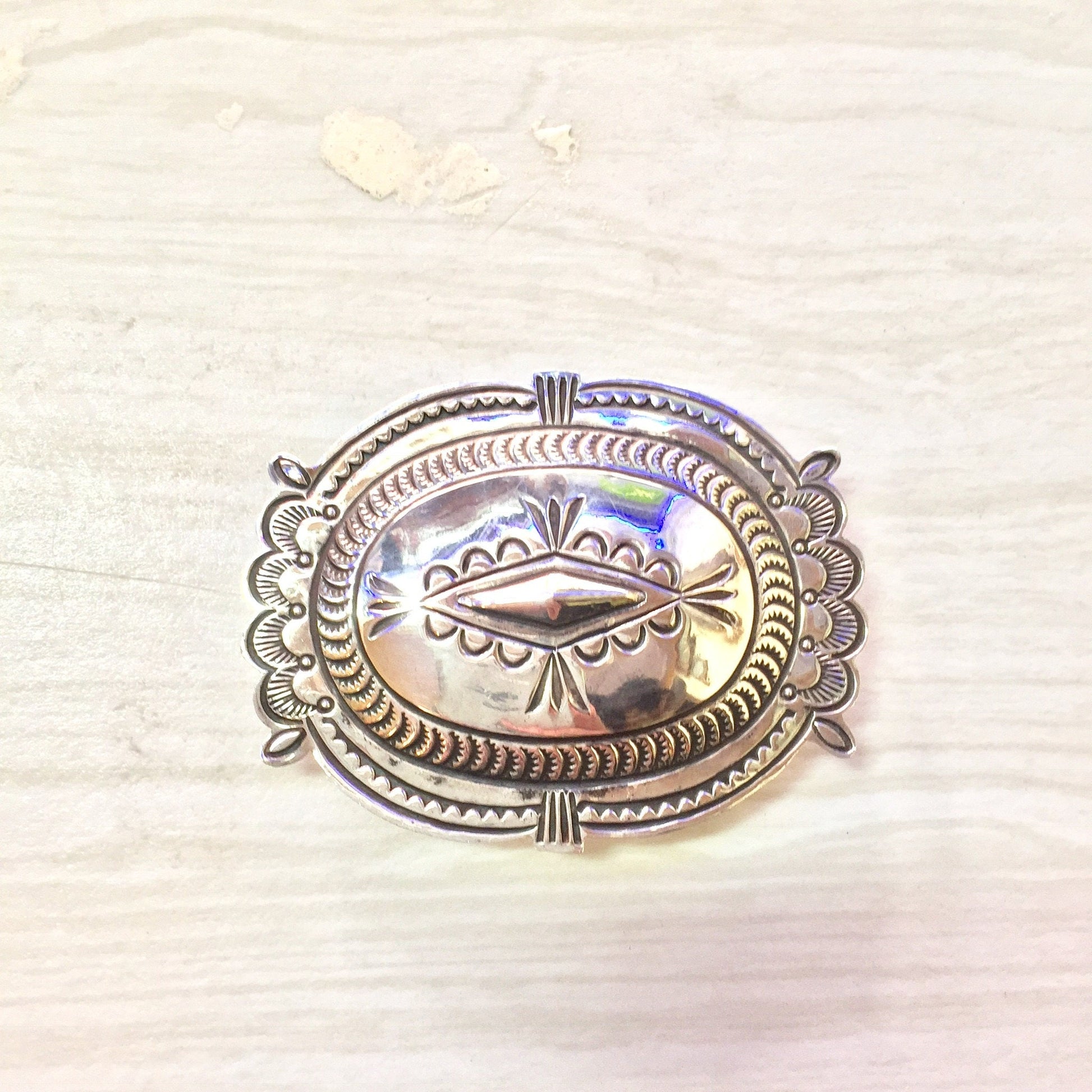 Sterling silver oval concho belt buckle with intricate etched designs and iridescent abalone shell inlay, in a southwestern vintage western style, on a light colored textured background.