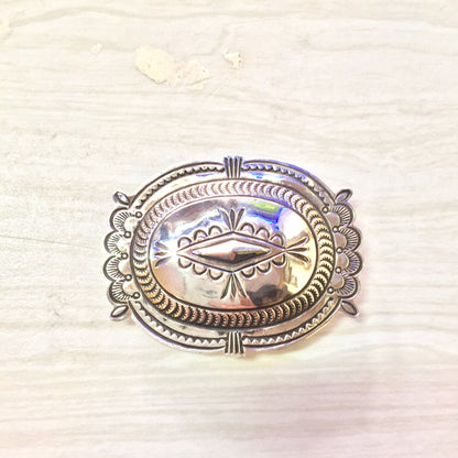 Sterling silver oval concho belt buckle with intricate etched designs and iridescent abalone shell inlay, in a southwestern vintage western style, on a light colored textured background.