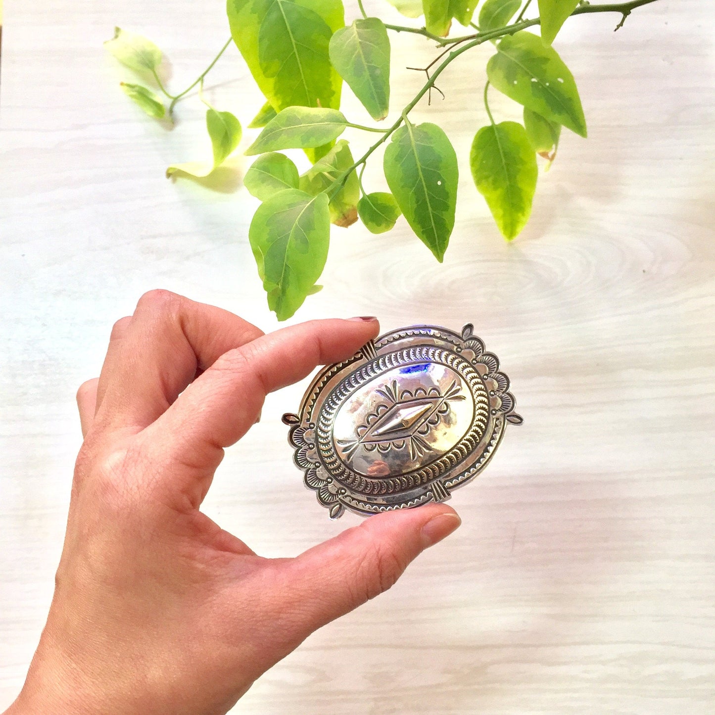 Vintage sterling silver concho belt buckle with southwestern design held in front of green leaves, western style accessories