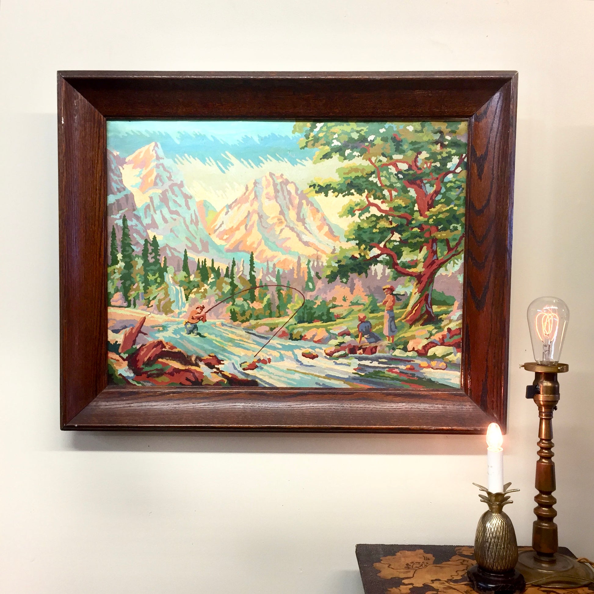 Large vintage paint by numbers oil painting depicting a mountain landscape with trees and a river, framed in a wooden frame, displayed on a wall above decorative items on a surface.