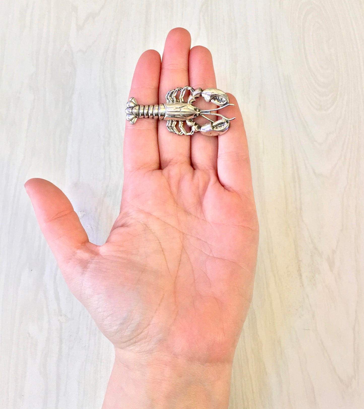 A hand holding a silver lobster brooch pin against a light wooden background.