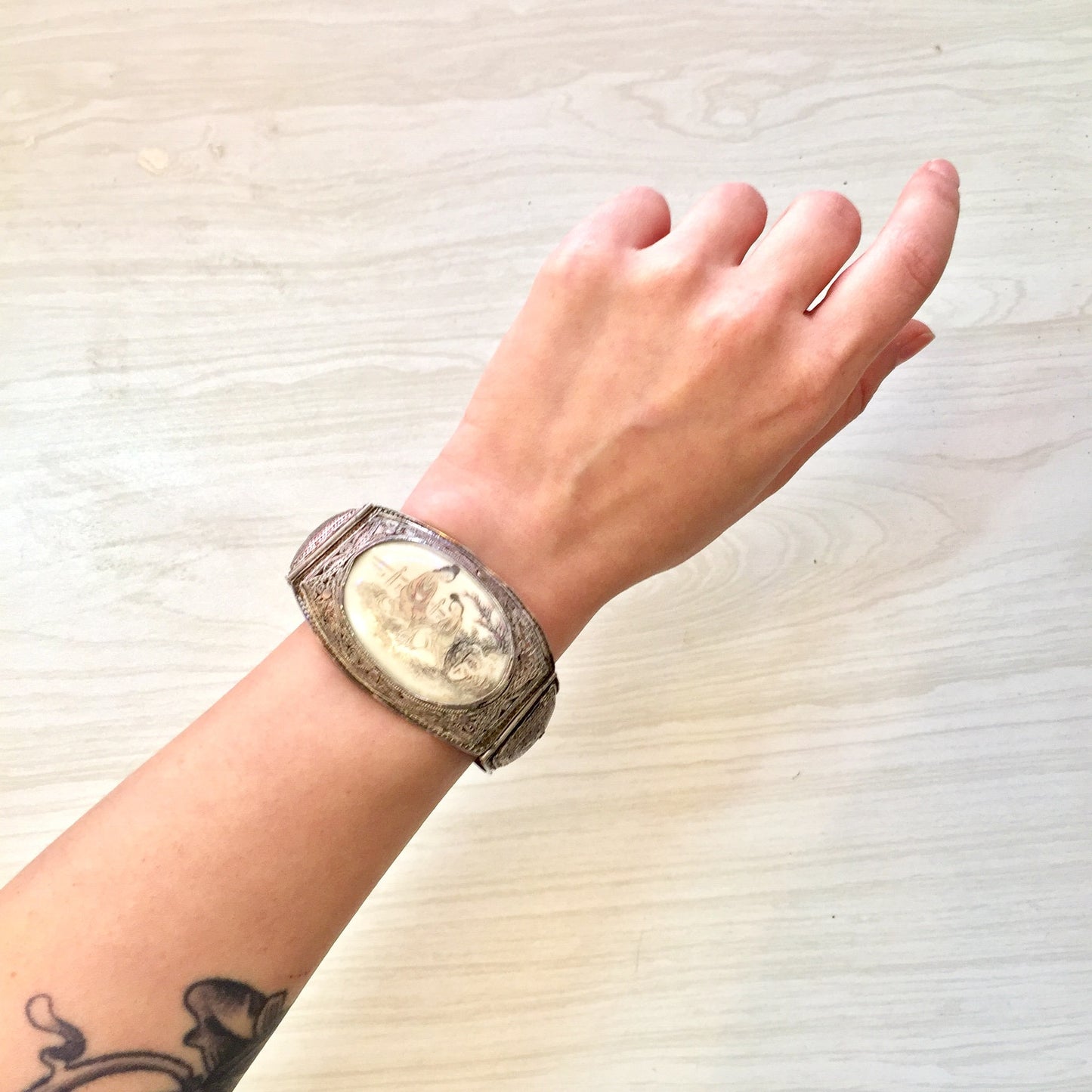 Vintage sterling silver hinged bracelet with intricately carved floral ivory or bone inlay on a tattooed wrist against a light wood background.