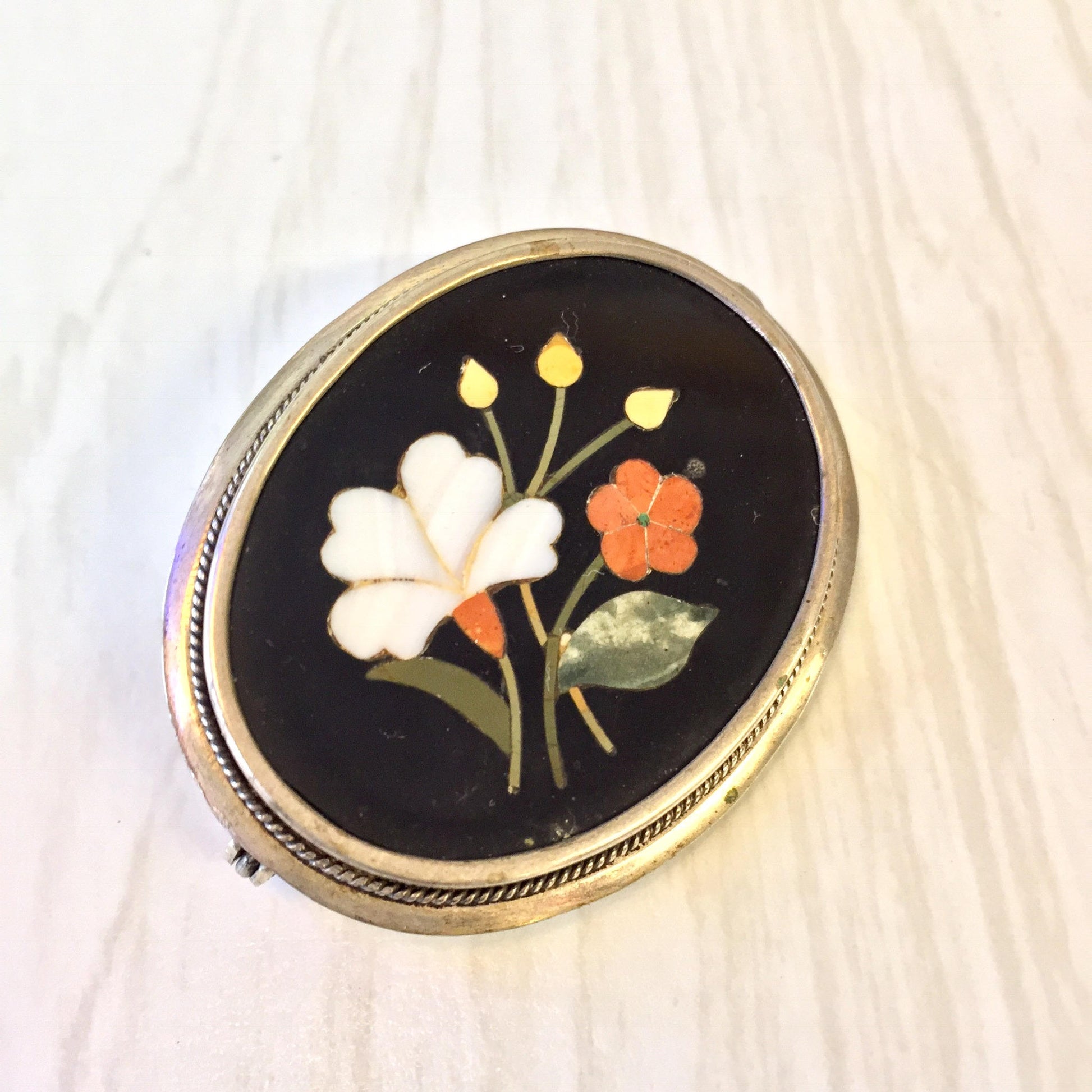 Sterling silver brooch with inlaid natural stone flowers including white and orange petals on a black background, set in a round gold-toned frame with textured border.