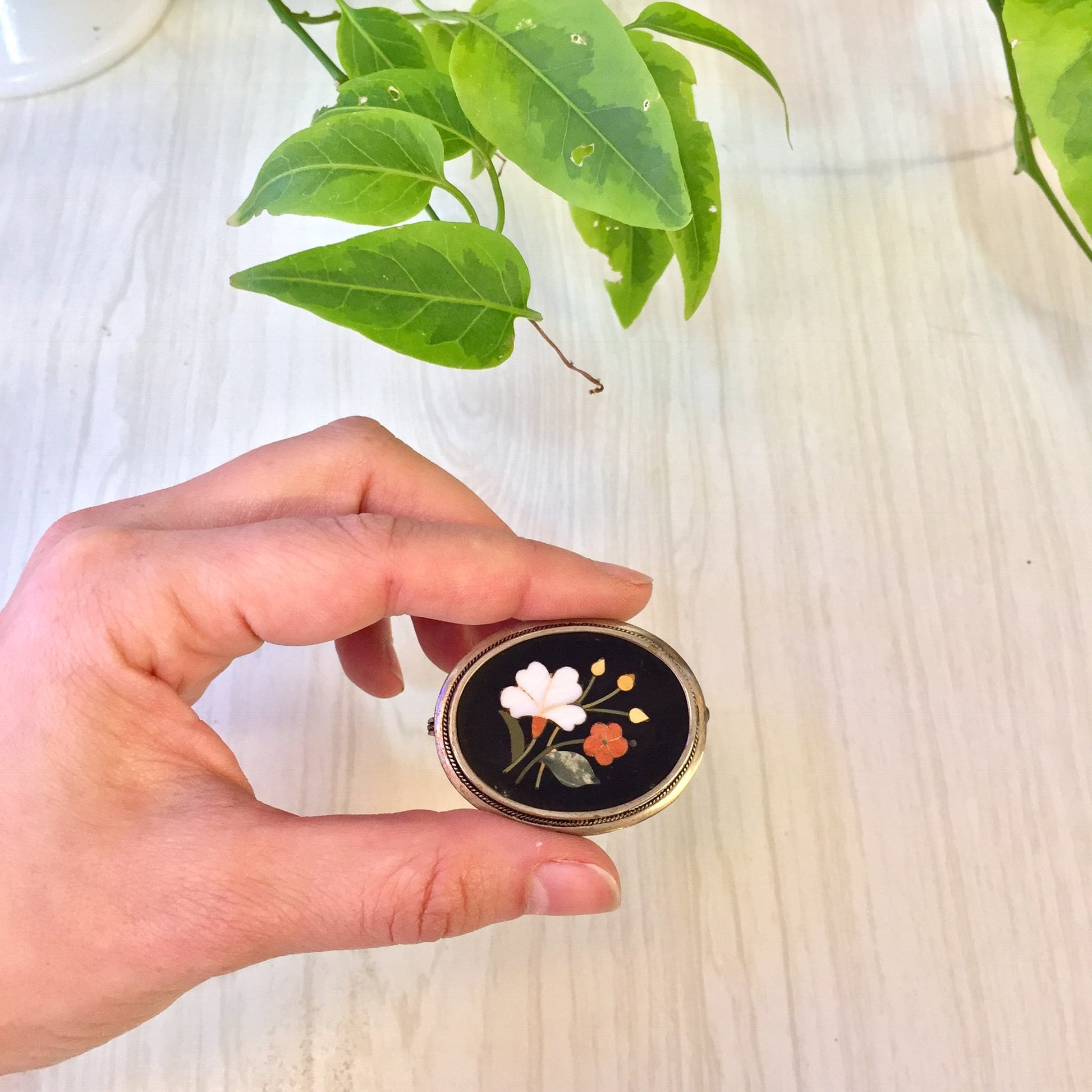 Vintage sterling silver brooch with inlaid natural stones in a floral design, held in front of plant leaves on a light wooden surface.