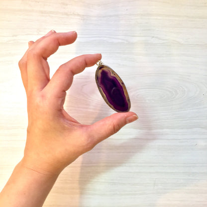 A hand holding a large sterling silver pendant with a vibrant purple agate stone against a light wooden background.