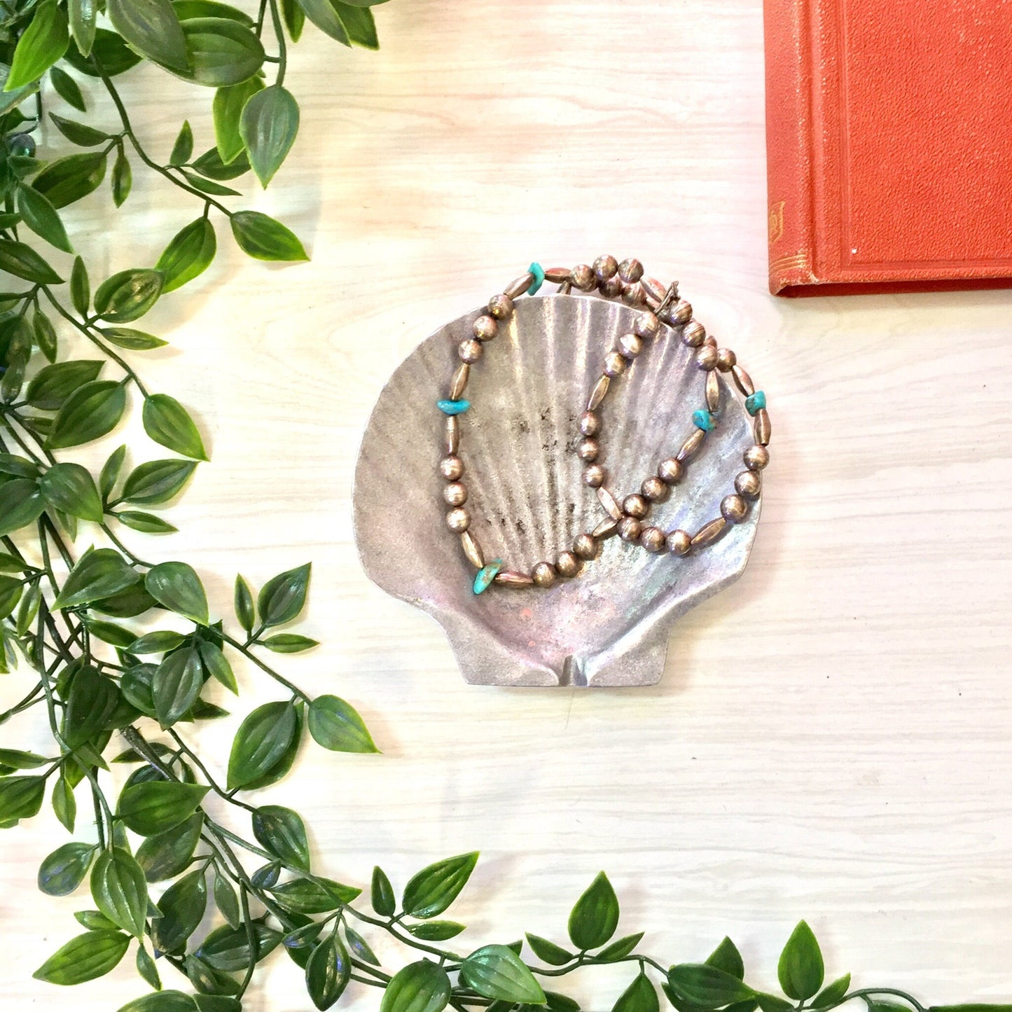 Vintage pewter seashell-shaped ash tray or jewelry dish with turquoise beads displayed on wooden surface with greenery and orange book.