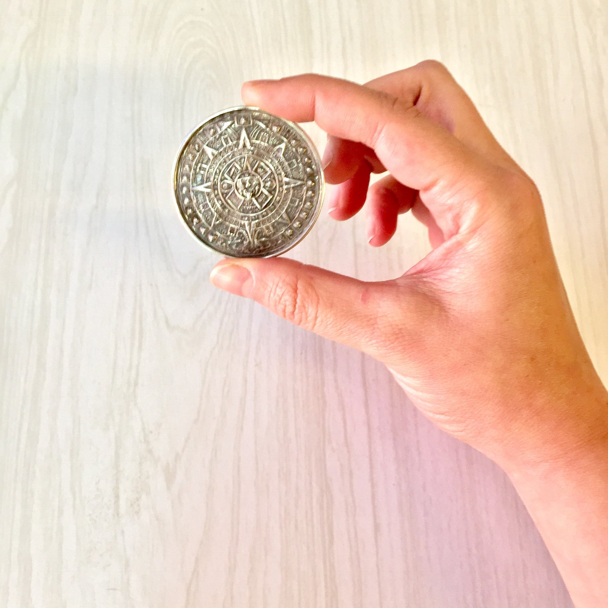 Hand holding a vintage sterling silver Mayan Aztec sun calendar pendant or brooch against a light wooden background, suitable as a unique holiday gift idea from the vintage jewelry collection.